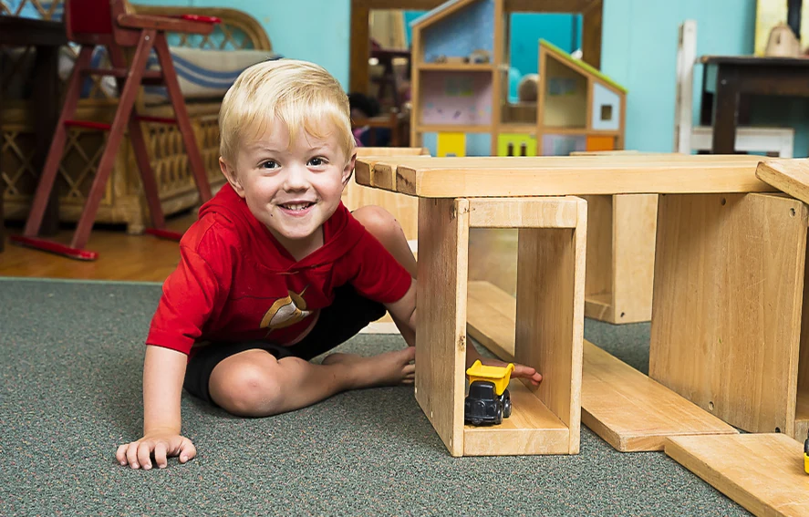 A smiling boy lying on the floor behind a wooden block tower